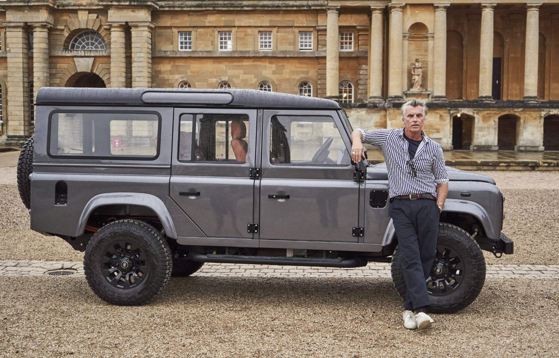 Photograph of a model dressed in Neem brand clothes, leaning against an electric Land Rover converted by Electrogenic. Blenheim Palace is in the background.