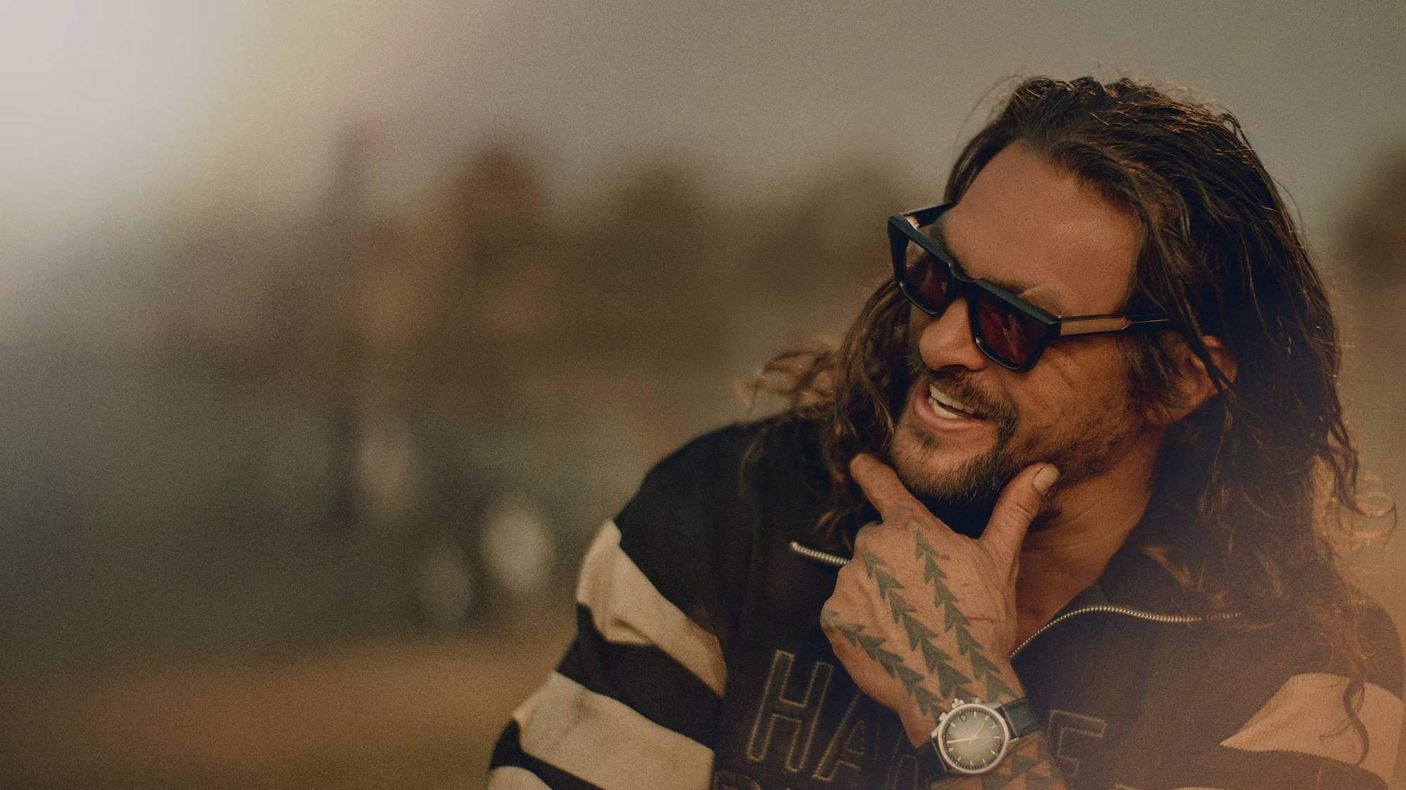 Photograph of Jason Mamoa smiling and wearing a pair of sunglasses. He is looking off-camera.