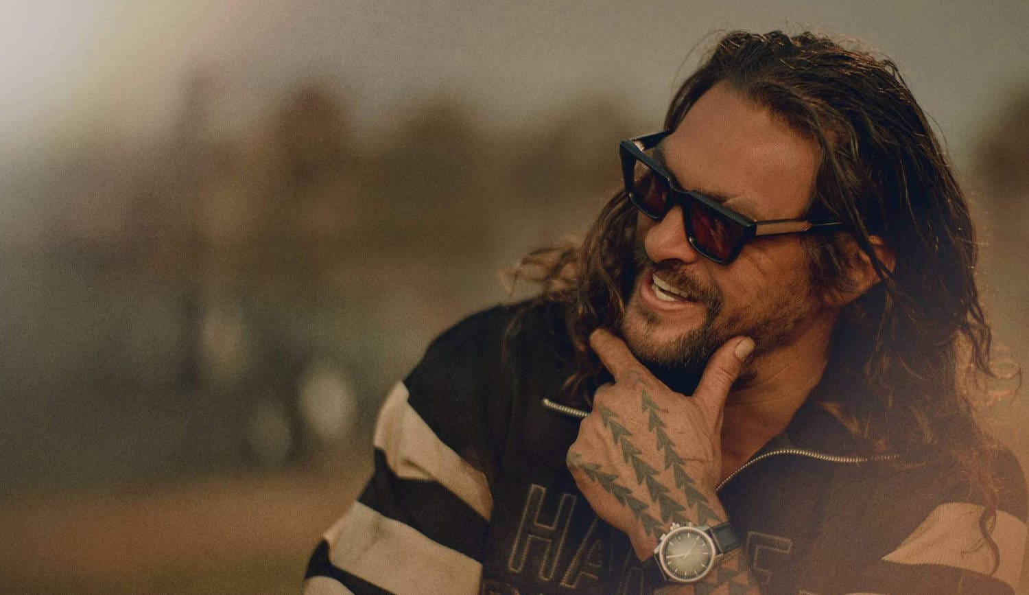 Photograph of Jason Mamoa smiling and wearing a pair of sunglasses. He is looking off-camera.